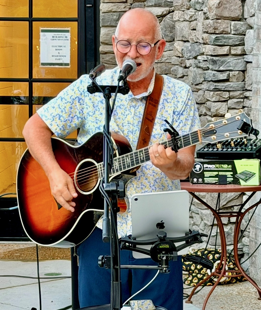Danny Dukes singing and playing acoustic guitar in a floral shirt beside a stone wall